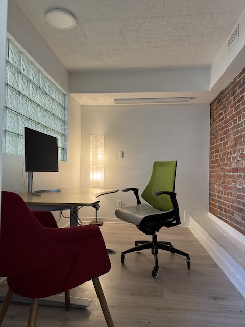 An office with desk and a chair, brick along one wall and a floor lamp providing soft light